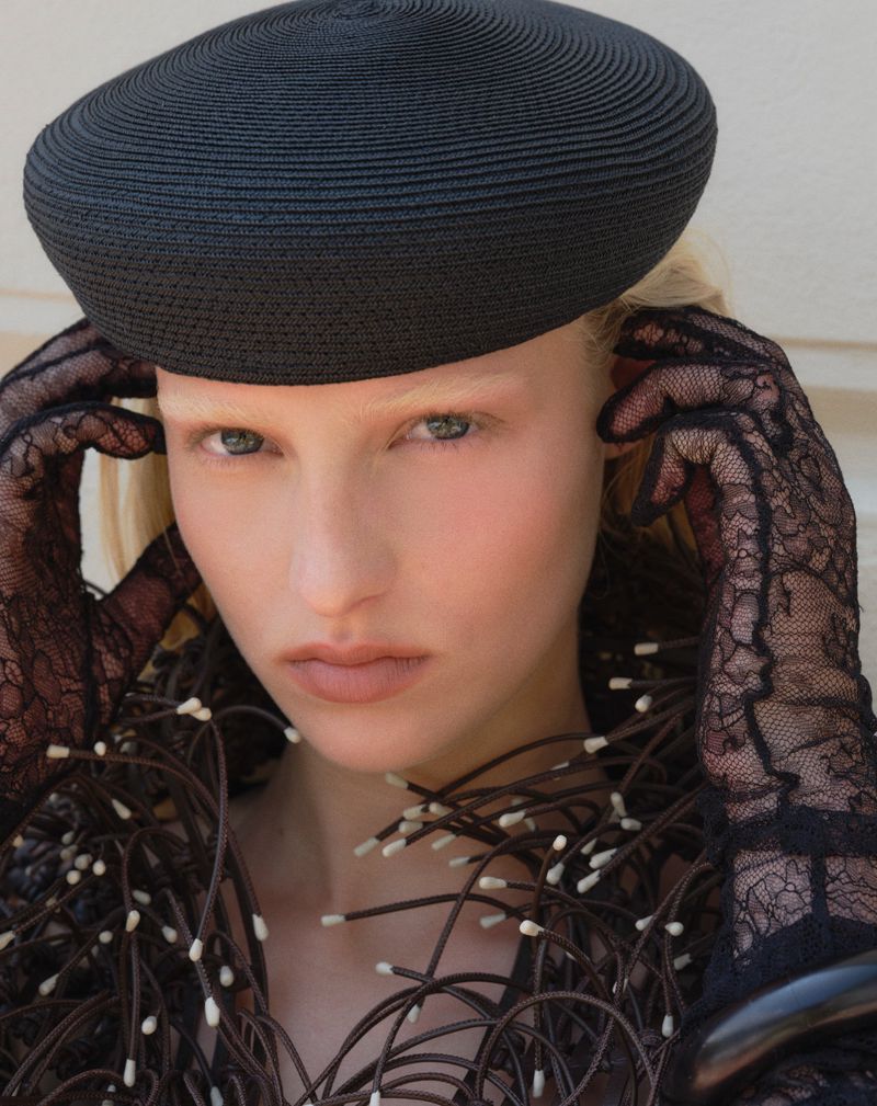 Close-up of a woman with light skin wearing a black hat and lace gloves, posing with decorative floral collar.