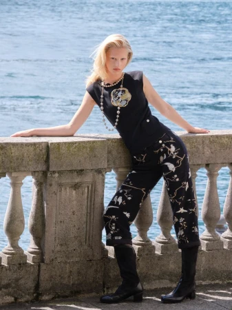 Woman in black top and floral pants posing on a stone ledge overlooking the sea in a fashion editorial shot, creating a striking visual.