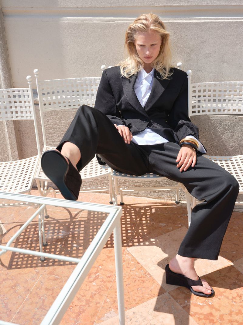Fashionable woman in blazer and trousers strikes a pose outdoors sitting on a white metal chair in the sunshine.