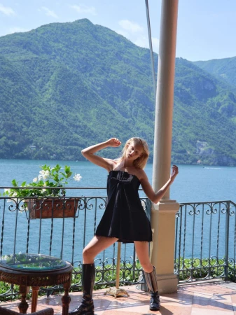 Young woman in black dress and boots poses on terrace with a lake and mountain backdrop on a sunny day. Balcony is ornate.