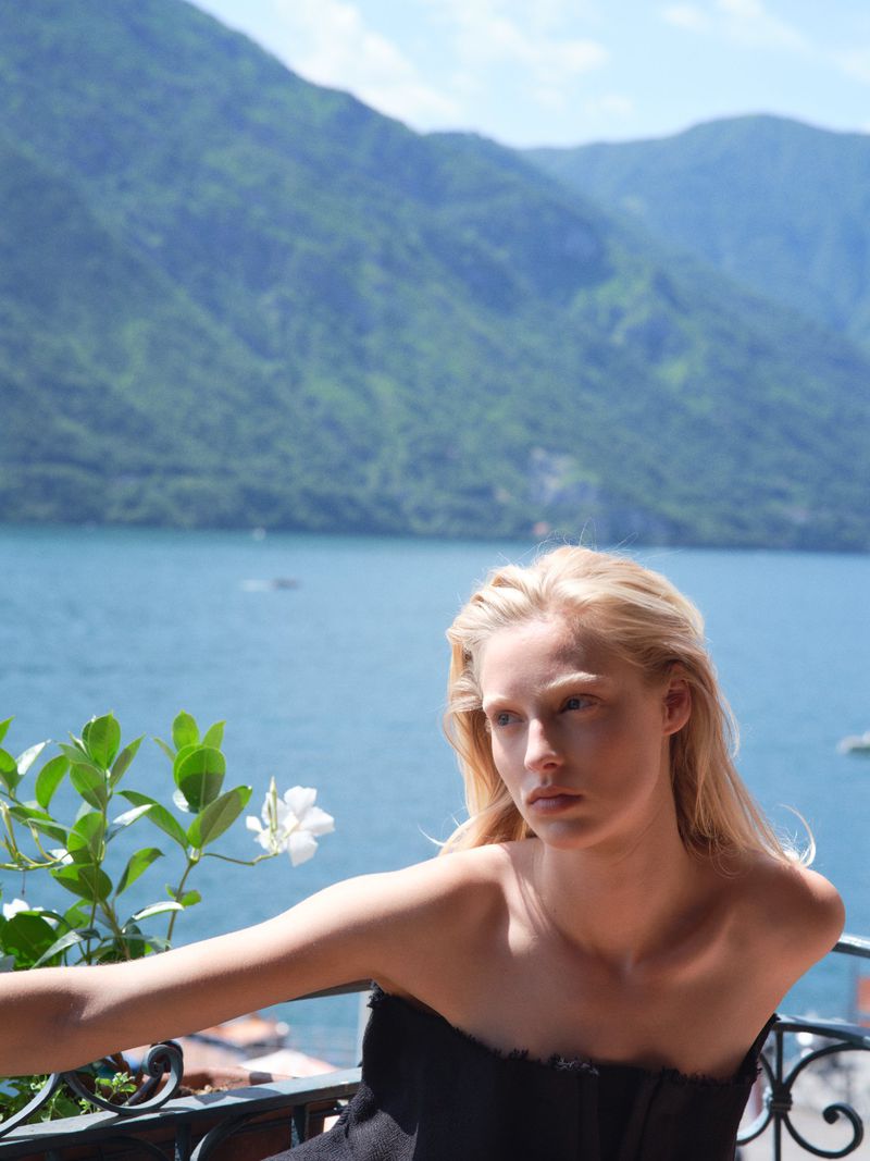Portrait of a blonde woman in a black strapless top on a balcony overlooking a lake and mountains on a sunny day.