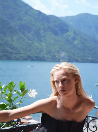 Portrait of a blonde woman in a black strapless top on a balcony overlooking a lake and mountains on a sunny day.