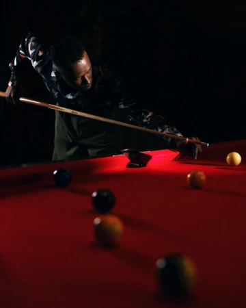 An African American man playing billiards in a dimly lit room, focused on aiming with a cue stick at the billiard balls on a red pool table.
