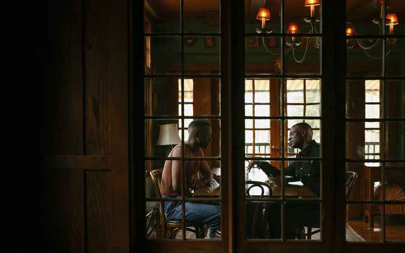 Two adult men in dark wood dining room engaged in conversation, visible through windowed doors, warm lighting, and soft focus background.