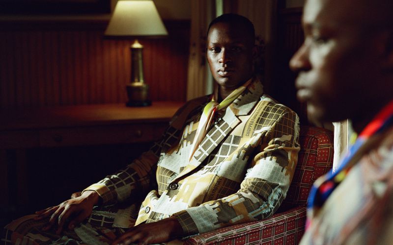 Two adult males in a dimly lit indoor scene. One man is in focus wearing a patterned suit. A table lamp is visible in the background.