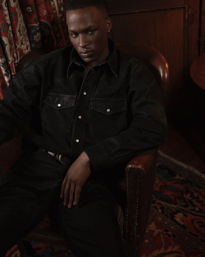 Portrait of an adult male in a button-down shirt sitting in a leather chair with a patterned rug and curtain backdrop.