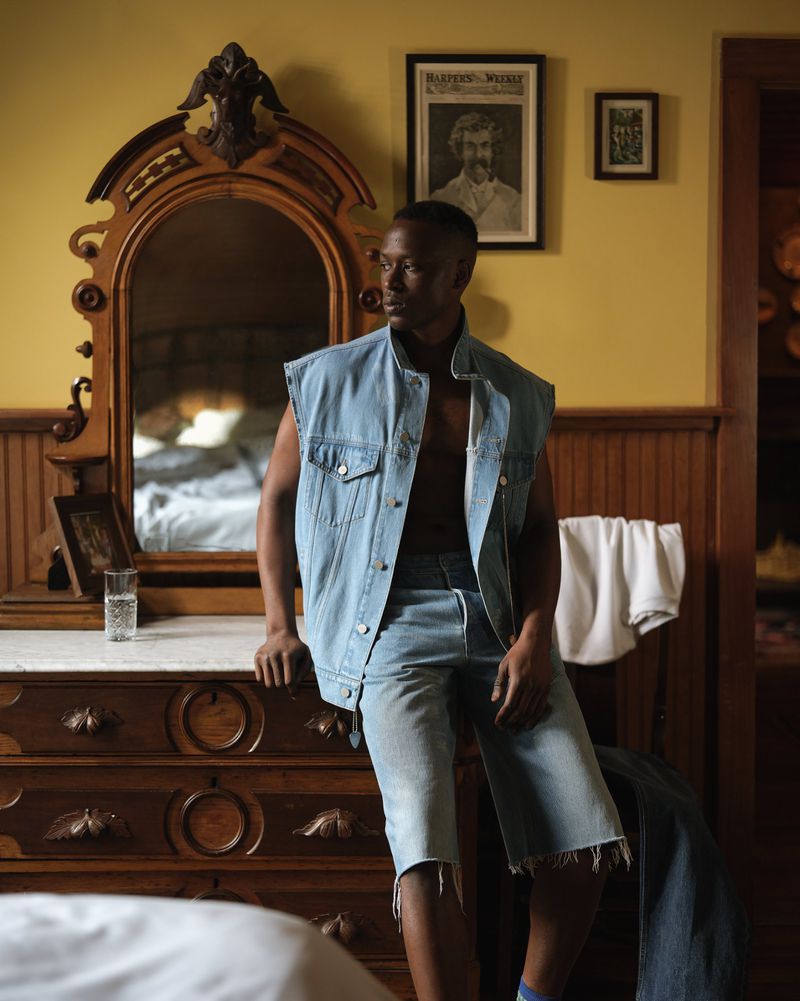 Stylish man in denim vest and shorts posing indoors with antique dresser and framed art, bathed in natural light.