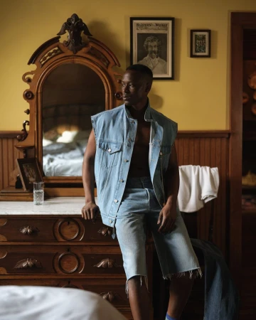 Stylish man in denim vest and shorts posing indoors with antique dresser and framed art, bathed in natural light.