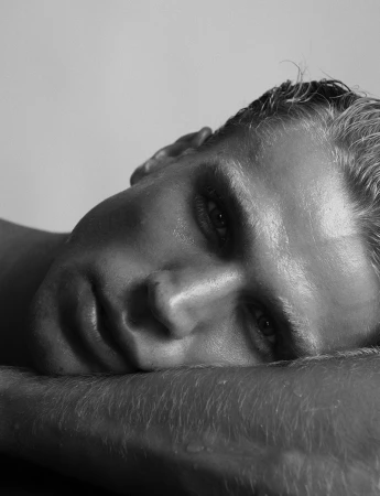 Black and white portrait of a sweaty adult male lying down with his head resting on his arm in a brightly lit studio setting.