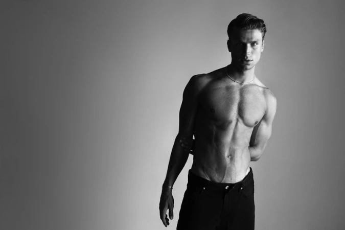 A bare-chested man stands in a studio with hands behind his back. He is wearing black pants. The background is a gradient grey.