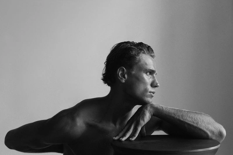 Black and white portrait of a shirtless adult male looking left with arms resting on a dark table against a plain background.
