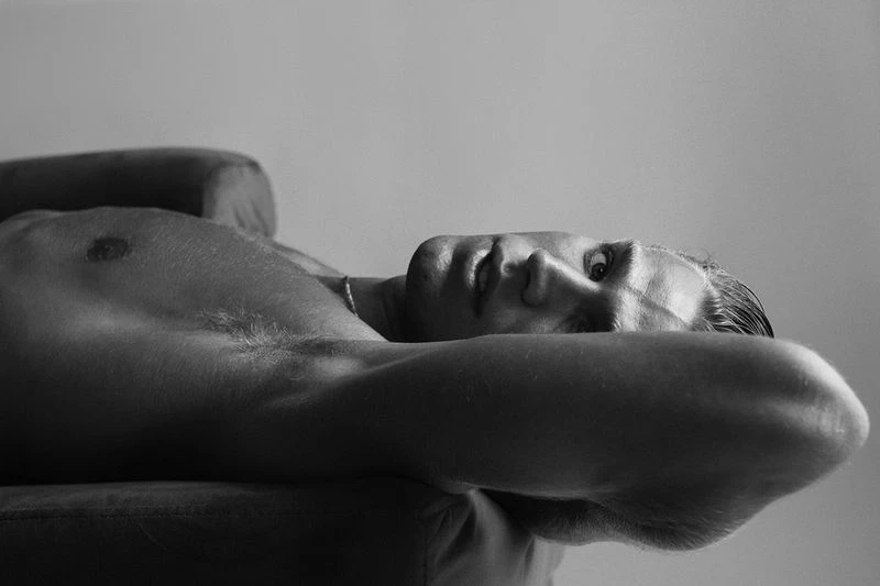 Black and white portrait of a fair-skinned adult male lying back on a sofa with arms crossed behind his head in a relaxed pose.
