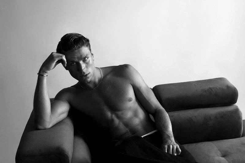 Black and white studio shot of a muscular adult male reclining on a couch, looking at the camera with a neutral expression.