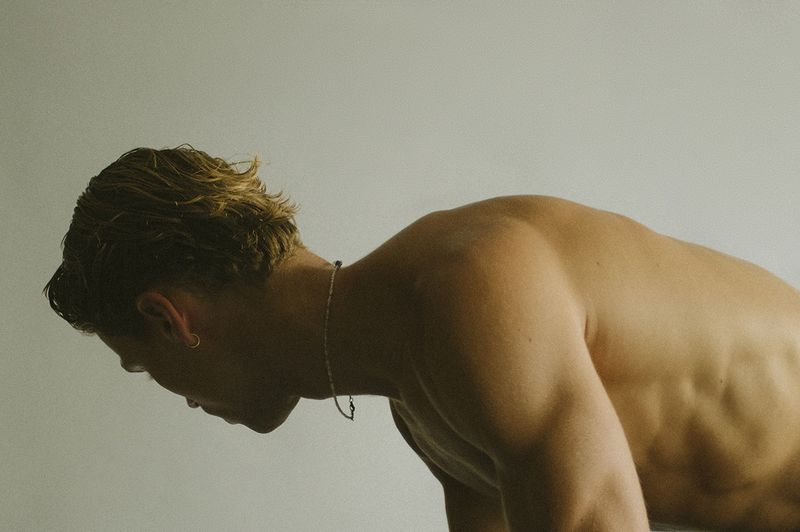Muscular topless man with a necklace bends forward in front of a solid grey background in a medium shot, revealing his strong upper back.