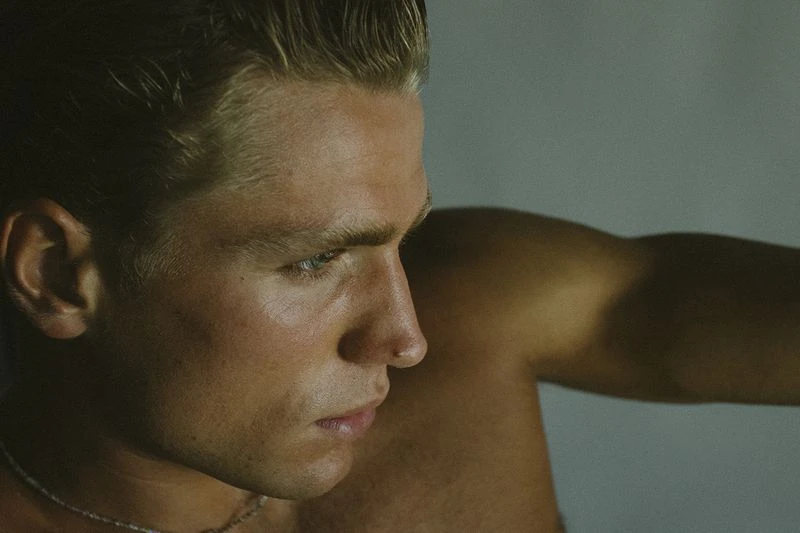 Close-up portrait of an adult male with blonde hair, fair skin, and a visible collarbone, set against a neutral, diffused background.