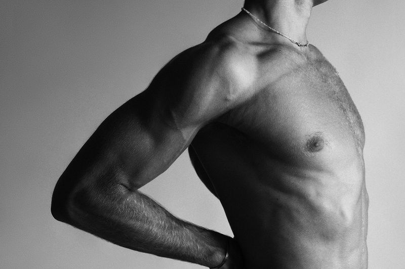 Monochrome close-up studio shot of a muscular adult man's bare chest and torso with arm raised against a plain grey background.