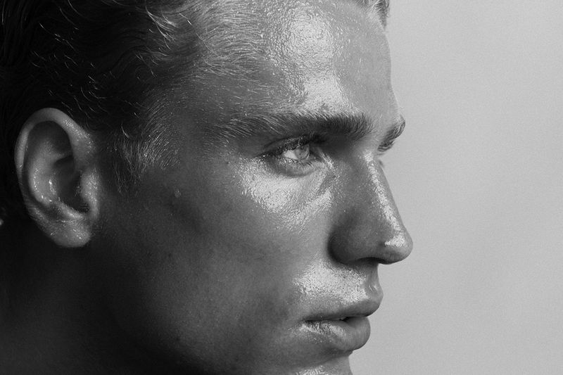 Close-up black and white portrait of a blonde male model with a wet, glistening face in a studio with soft, diffused lighting.