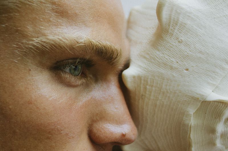 Close-up profile of a fair-skinned adult male's face with an aqua eye next to a textured, organic object. Soft, warm lighting.