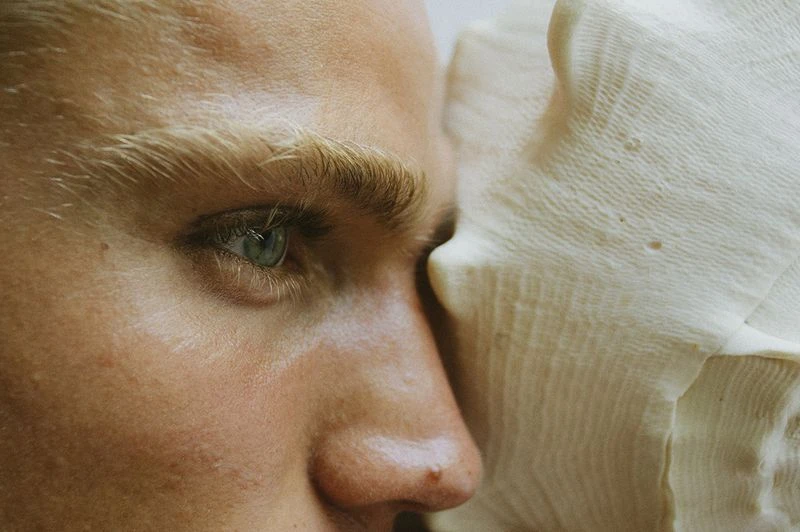 Close-up profile of a fair-skinned adult male's face with an aqua eye next to a textured, organic object. Soft, warm lighting.
