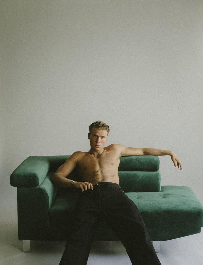 Shirtless man with blond hair sits on a green velvet couch in a bright indoor setting, striking a casual pose with a neutral facial expression.