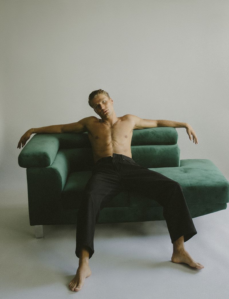A shirtless man with dark jeans sits on a dark green velvet sofa in a brightly lit interior, posing for a fashion shot.