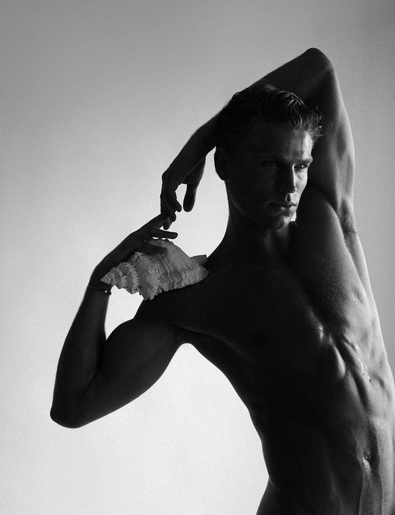 Black and white portrait of shirtless adult male model with arms raised, holding a shell against a plain background in studio lighting.