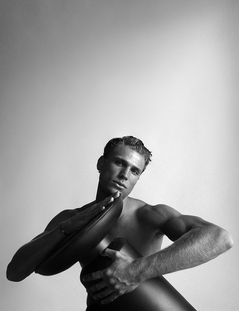 Black and white portrait of a shirtless adult male with short hair holding an oval object, posing against a plain light background.
