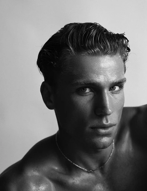 Monochrome portrait of a muscular adult male with slicked-back hair and a silver necklace, captured in a close-up studio setting.