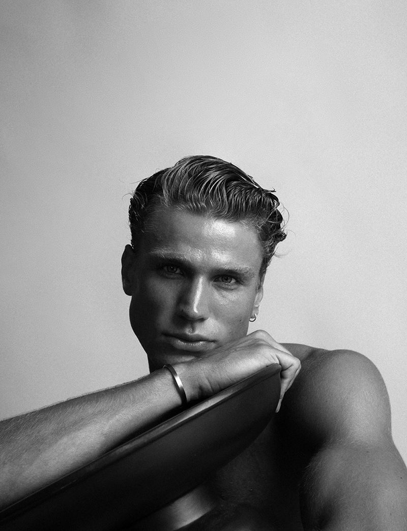Monochrome portrait of a fair-skinned adult male with slicked-back hair, looking at the camera, resting his arm on a smooth surface.