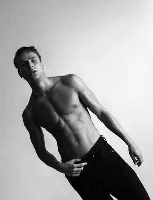 Shirtless man wearing dark trousers and a thin chain necklace posing against a plain white studio background.