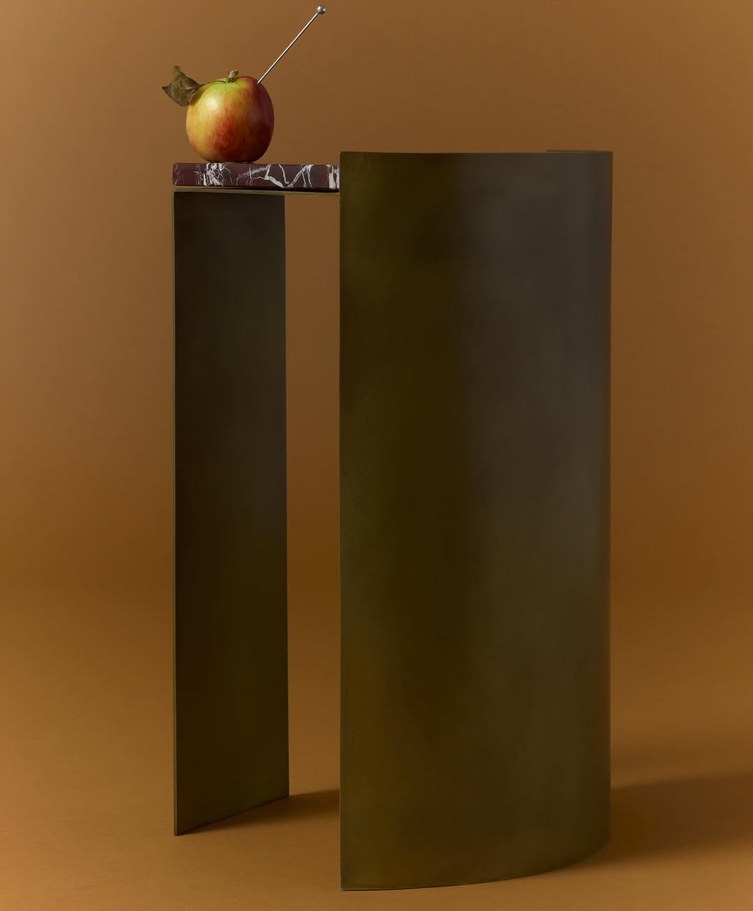 Still life featuring a red apple with a metal skewer on a geometric pedestal table against a tan studio backdrop.