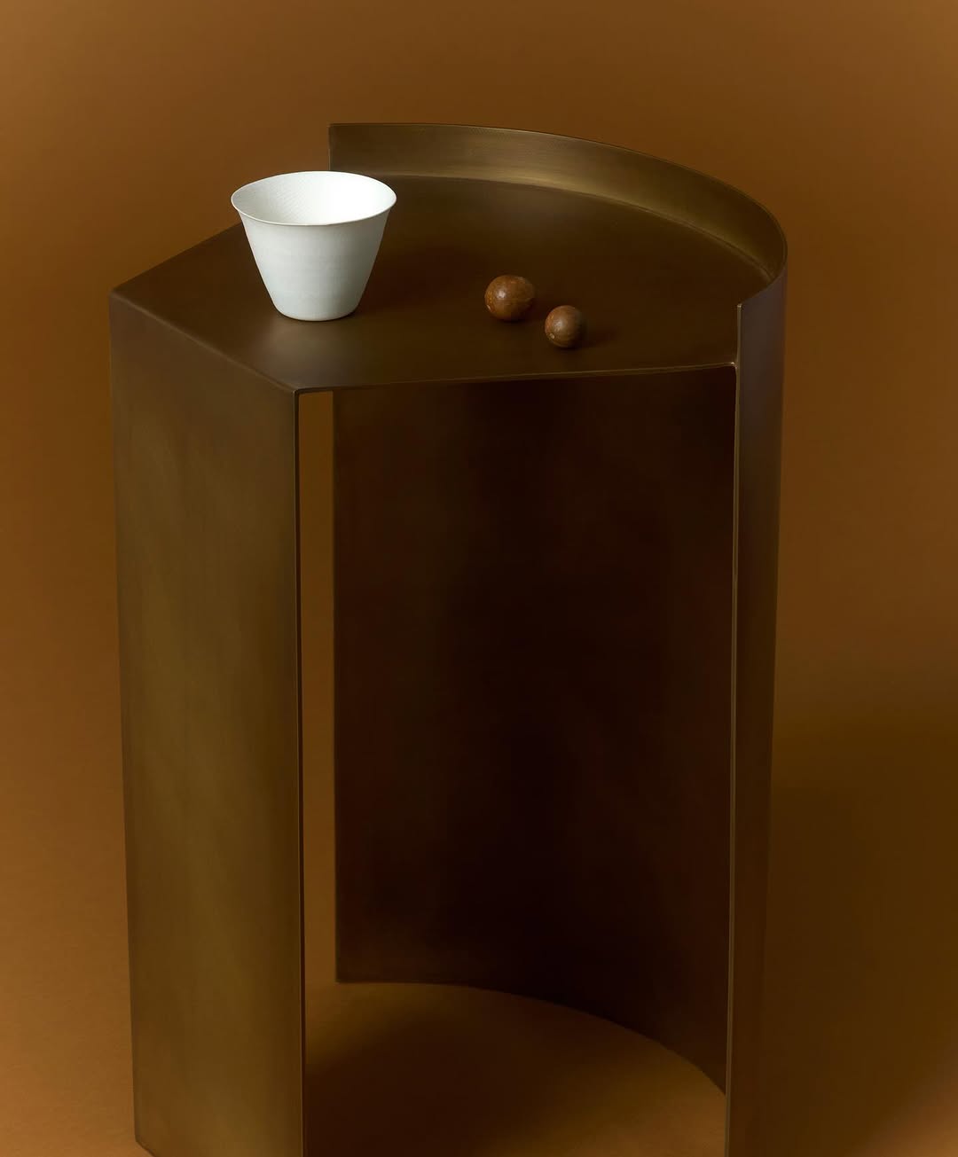 A brass end table with a white ceramic bowl and two brown spheres on top against a solid brown background in a studio setting.