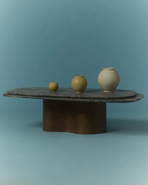 Three vases sit on a stone oval table with a sculptural metal base, all against a solid blue background in a studio setting.