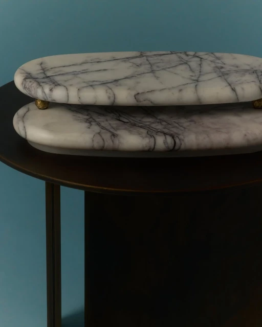 Two oval marble trays with brass feet stacked on a dark brown side table against a teal background in a brightly lit studio.