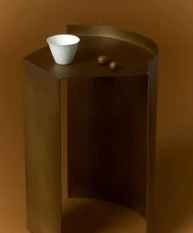 Minimalist bronze curved side table holding a white ceramic cup and two small round objects against brown background.