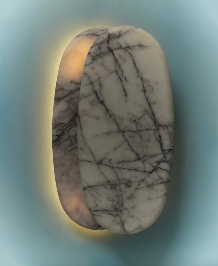 Sculptural oval marble wall sconce emitting a warm backglow against a pale blue wall for Just Adele's Oro Collection
