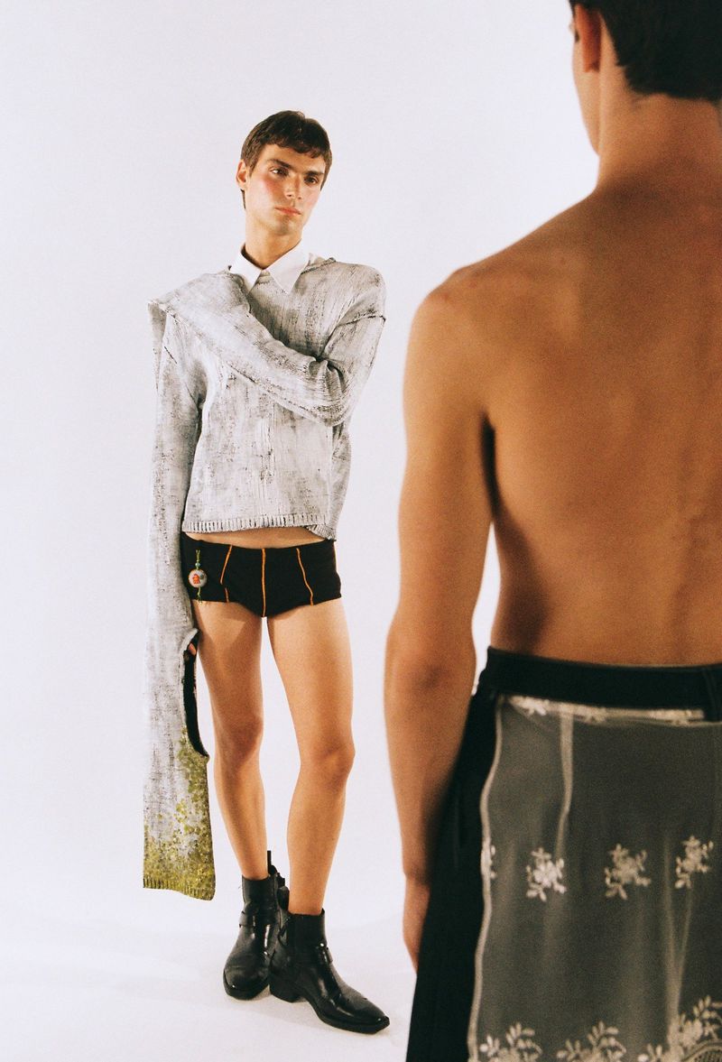 Two young adult men pose in a bright photo studio. One wears an oversized sweater, boxer shorts, and boots, while the other is shirtless with a wrap.
