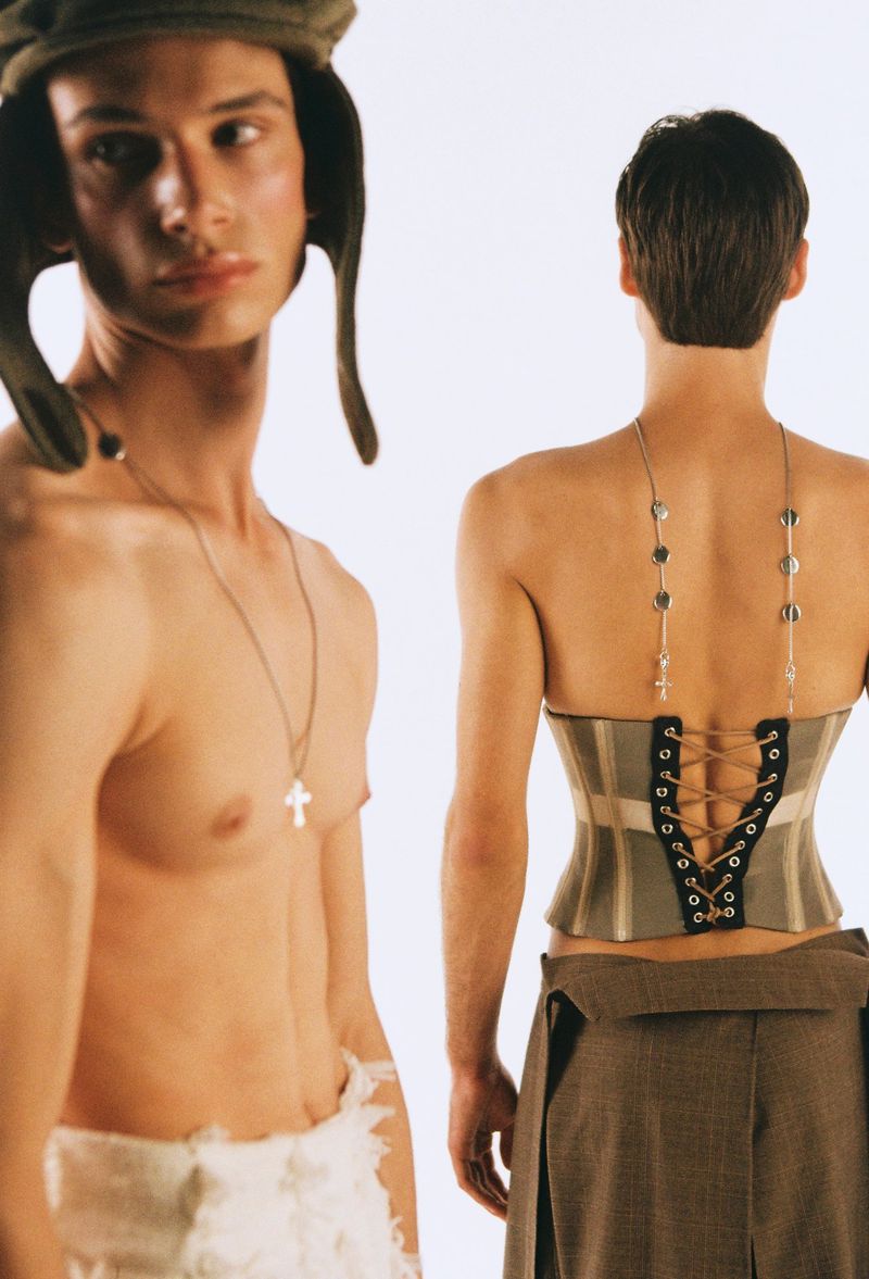 Two shirtless, young adult men pose in a photo studio. One wears a cap; the other wears a corset and skirt.