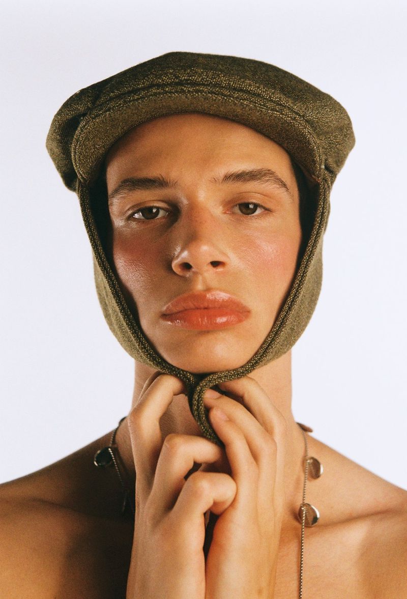 Close-up of a young adult with tan skin, wearing an olive-green cap and silver necklace, with both hands near their face in a studio setting.