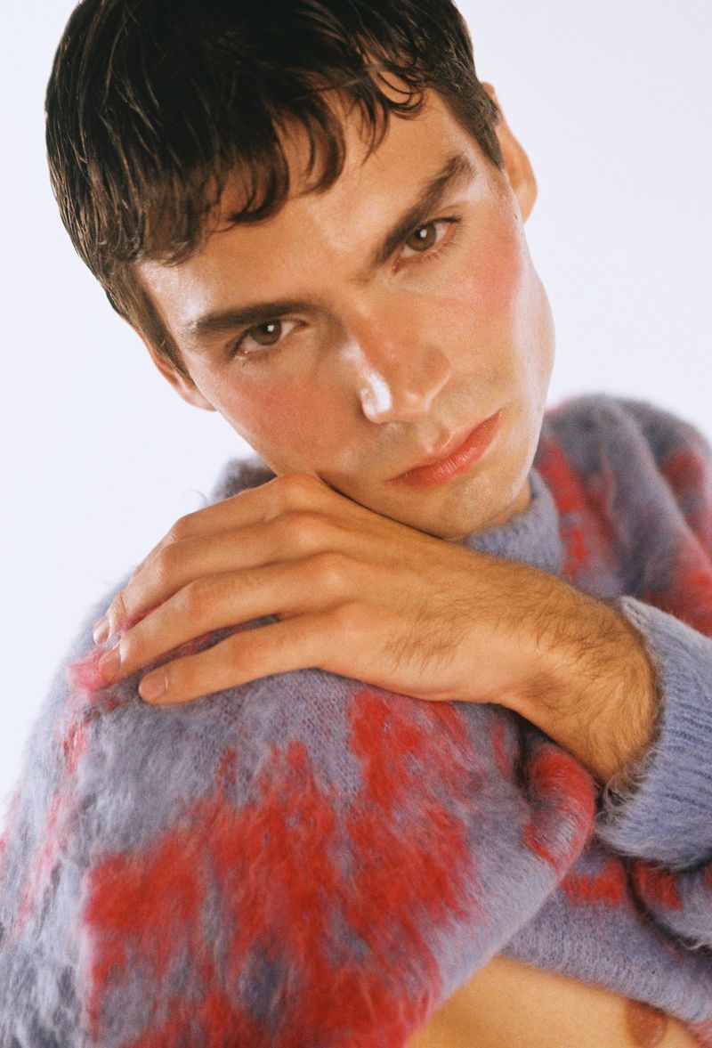 Portrait of a young adult man with light skin and brown hair wearing a patterned sweater, gazing at the camera in a studio setting.