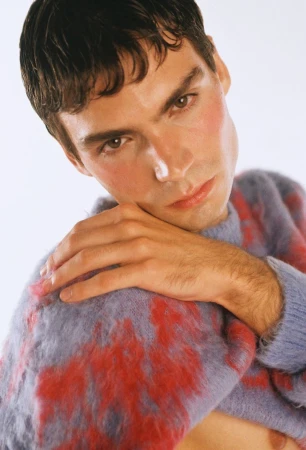 Portrait of a young adult man with light skin and brown hair wearing a patterned sweater, gazing at the camera in a studio setting.