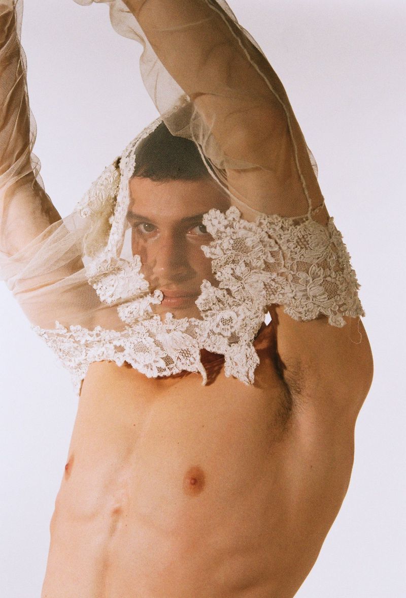 Portrait of a shirtless adult male with dark hair, partially obscured by sheer white lace fabric, posing against a stark white background.