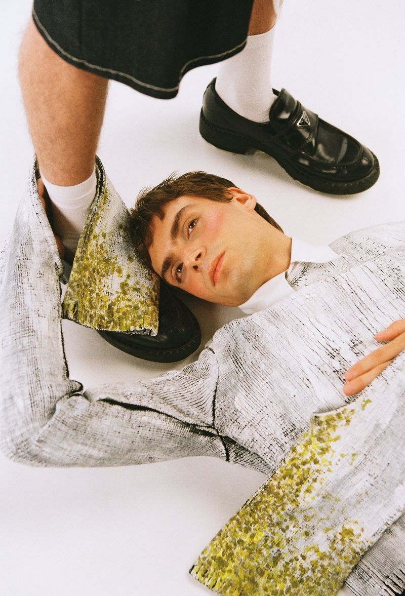 Two young adult men strike a pose in a studio setting. One man rests his head on the foot of the other, wearing casual fashion.