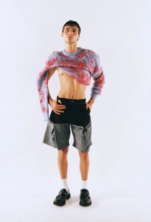 Fashion shot of a slender man in grey shorts and a colorful cropped sweater standing in a white photo studio with soft, even lighting.