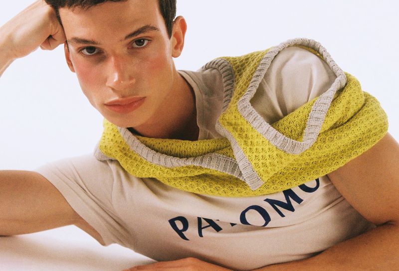 A young man lies on a white surface wearing a t-shirt and a yellow knit scarf-like top. He's gazing into the camera with a slight smile.