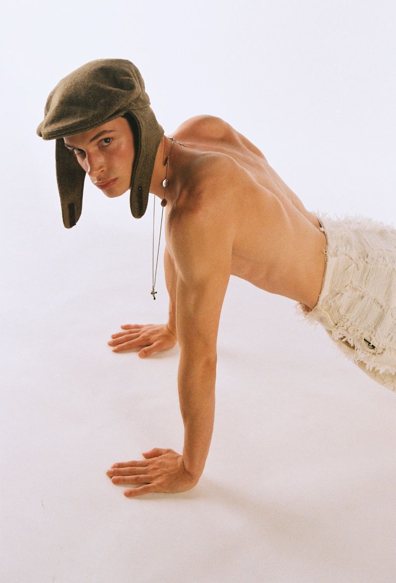 Male model poses in push-up position wearing a newsboy cap and jewelry against a white background, creating a unique studio portrait.