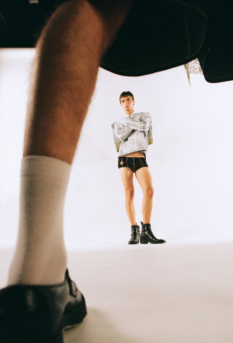 Man in fashion clothing poses in a studio, viewed from low angle perspective, with partial figure of another person in foreground.
