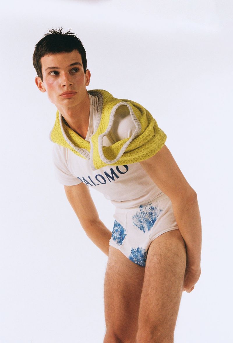 A slender young adult poses in a white t-shirt, patterned briefs, and a knitted yellow shoulder covering against a white background.