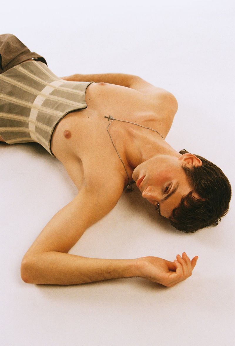 A young adult man with dark hair and a cross necklace lies on a white surface, wearing a grey corset with brown pants, artful and subdued.
