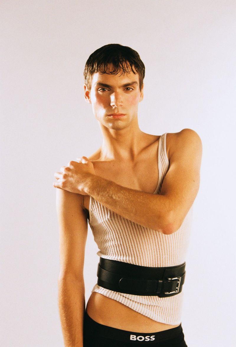 Adult male model in a photo studio wearing underwear and a tank top with an oversized leather belt and light makeup, looking at the camera.
